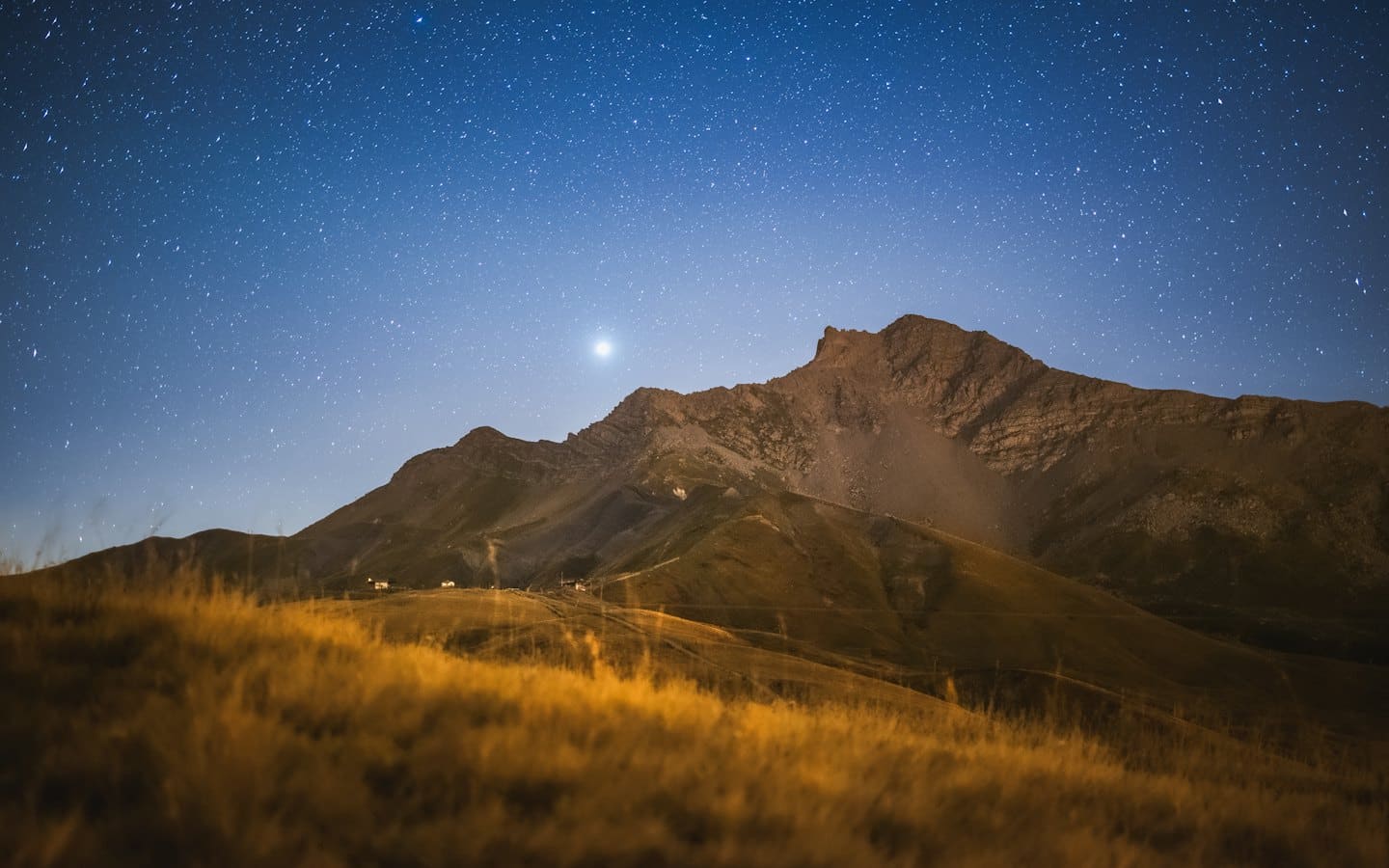 Işıklı tepeler ve silüetli dağların üzerinde yıldızlı gece gökyüzü.